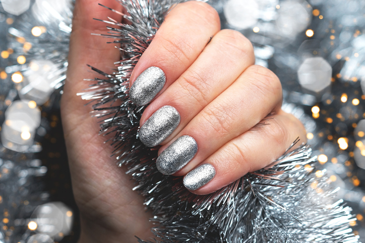 Female hand with beautiful holiday manicure - silver glittered nails on Christmas tinsel background with blurred lights